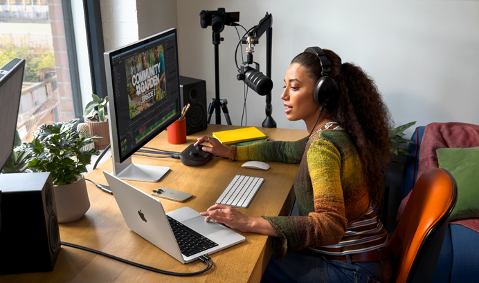 En kvinde arbejder hjemme ved sit skrivebord på MacBook Pro med hovedtelefoner på og to eksterne skærme tilsluttet, mens hun taler ind i en mikrofon