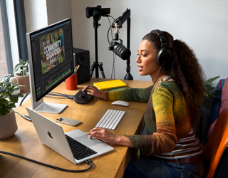 En kvinne jobber ved skrivebordet sitt hjemme med MacBook Pro koblet til to eksterne skjermer. Hun har på seg hodetelefoner og snakker i en mikrofon.