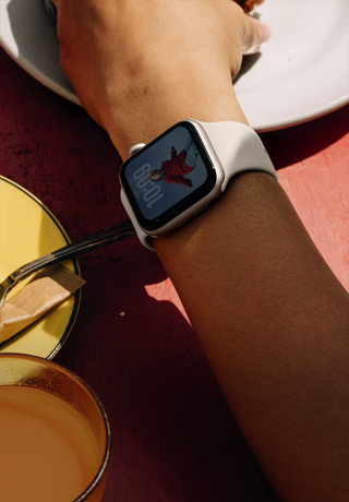 Henkilön käsivarsi kurottamassa ottamaan aamiaista, ranteessa on Apple Watch SE 3, tähtivalkea alumiinikuori, aina päällä oleva näyttö, henkilön muotokuva kellotaulussa, tähtivalkea urheiluranneke